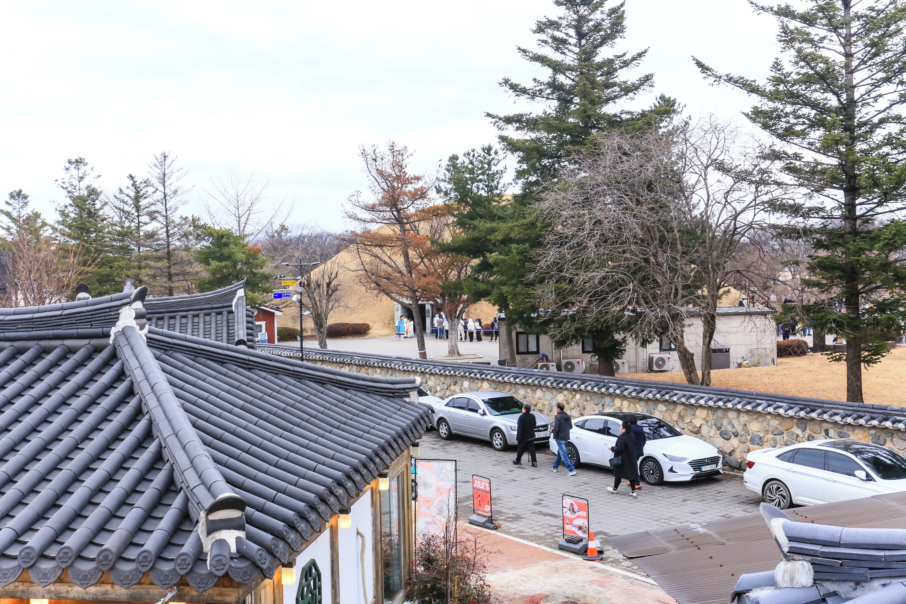 A traditional building with a tiled roof, a stone wall, and a paved area with parked cars.