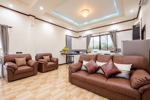 Een ruime woonkamer met een bruine leren bank, twee fauteuils, een eethoek en een keuken op de achtergrond, plus een groot raam voor natuurlijk licht.