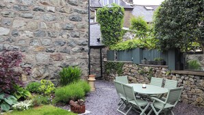 Un mur de pierres, une terrasse en gravier, une table verte avec des chaises et diverses plantes.