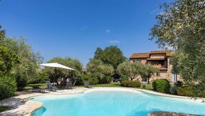 Una piscina de borde curvo, rodeada por una terraza de piedra. Hay un área de patio con sillas y una sombrilla.