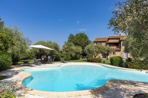Ein Pool mit geschwungener Kante, umgeben von einer Steindeck. Eine Terrasse mit Stühlen und Sonnenschirm.