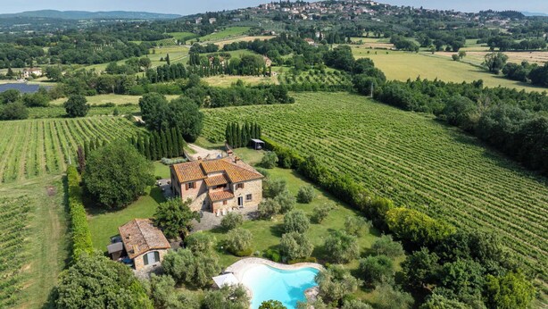 Una villa con piscina rodeada de viñedos, ubicada en un paisaje montañoso con un pueblo a lo lejos.