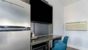 Eine kompakte Küchenzeile mit integriertem Kühlschrank, einer Mikrowelle und einem kleinen Backofen. Ein Flachbildfernseher ist über einer grauen Arbeitsplatte angebracht. Ein blauer Stuhl steht rechts.