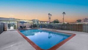 Una piscina con cerramiento de madera, una zona de estar cubierta y un edificio al fondo.