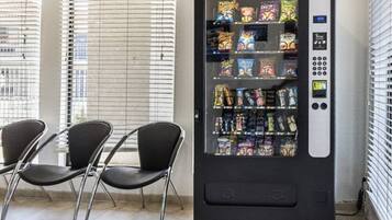 A vending machine with snacks and a payment interface, located in a room with chairs and blinds.
