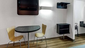 Quarto com televisão de tela plana na parede, uma pequena mesa com duas cadeiras, uma estante preta e um aspirador de pó.