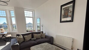 Ein modernes Wohnzimmer mit einem grauen Sofa, einem gemusterten Pouf und einem gerahmten Bild an der Wand.