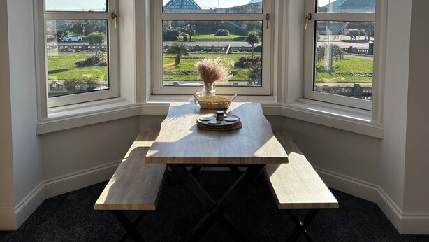 Ein Eckzimmer mit drei Erkerfenstern, einem Holztisch mit einer Vase und dekorativen Tellern sowie Blick auf eine grüne Landschaft und entfernte Berge.