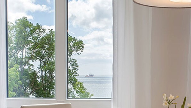 Habitación con un gran ventanal que ofrece vistas al mar y a los árboles, una silla blanca y una lámpara de pie.