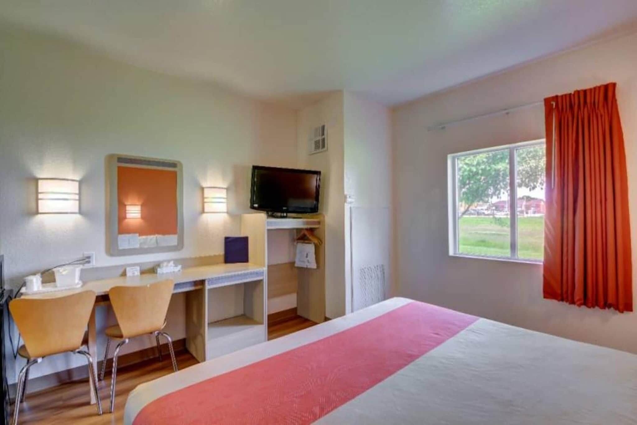 A hotel room with a bed, desk, chairs, a television, and a window with curtains.