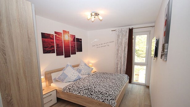 Une chambre à coucher avec un lit en bois, une table de chevet, une lampe fixée au mur et une fenêtre avec des rideaux.