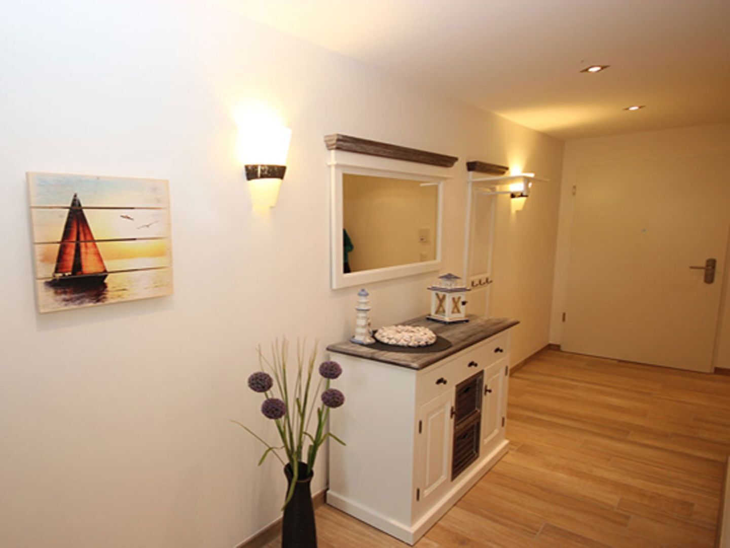 A hallway with a white cabinet, a vase with purple flowers, a painting of a sailboat, and wall sconces.