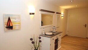 Un couloir avec une armoire blanche, un vase contenant des fleurs violettes, un tableau représentant un voilier et des appliques murales.