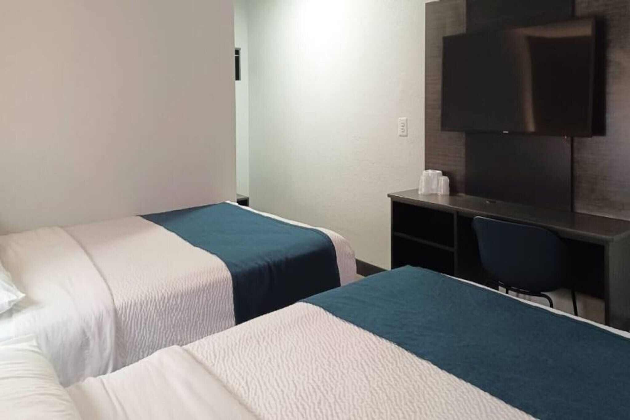 Quarto de hotel com duas camas, uma escrivaninha, uma cadeira e uma televisão de tela plana na parede.