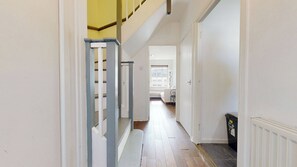 Ein schmaler Flur mit Treppe links, Heizkörper rechts und einer Tür am Ende, die zu einem Zimmer mit Bett und Fenster führt.