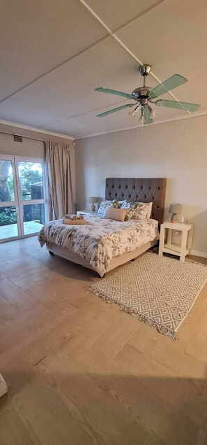 Une chambre à coucher avec un lit, un ventilateur de plafond, une table de chevet, un tapis à motifs et une vue sur l’extérieur, visible à travers une porte vitrée.