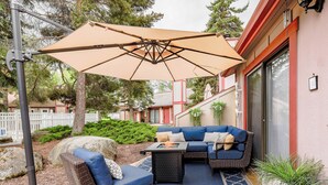 Et udendørs siddeområde med stor parasol, fletmøbler og udsigt til en bygning med rødlakeret dør.