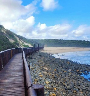Ein hölzerner Steg führt zu einem Strand mit felsigem Ufer und blauem Himmel.