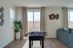 Ein Wohnzimmer mit einem Tischfußballtisch, einem grauen Sofa, einer Topfpflanze und einer Glasschiebetür, die auf einen Balkon führt.