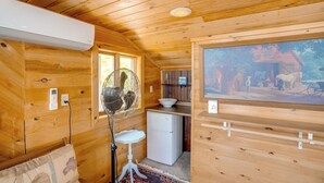 Interior de una cabaña de madera con un ventilador, un refrigerador pequeño, un lavamanos y un cuadro en la pared.