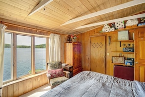 A wooden cabin bedroom with a large window, a bed, a chair, and a wardrobe.