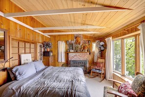 Une chambre dans une cabane en bois, avec un lit, une cheminée, un fauteuil à bascule et une grande fenêtre.