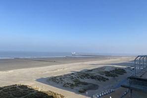 Ein Strand mit einem Steg, der ins Meer führt, einem sandigen Bereich mit Dünen und einem Gebäude mit einer Treppe.