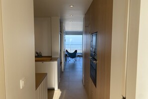 Ein schmaler Flur mit einer Küchenzeile links, einer Essecke mit blauem Stuhl in der Mitte und Blick auf das Meer durch große Fenster.