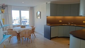 Eine moderne Küche mit einer gewölbten Kücheninsel, einem Essbereich mit rundem Tisch und Korbstühlen sowie ein Blick durch ein großes Fenster.
