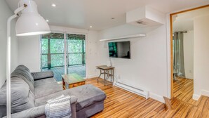 Een moderne woonkamer met een grijze bank, een houten salontafel, een witte vloerlamp, een flatscreen-tv aan de muur en grote ramen met uitzicht op groen.