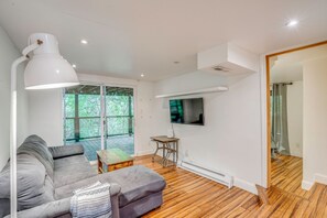 Ein modernes Wohnzimmer mit grauem Sofa, einem Holztisch, einer weißen Stehlampe, einem an der Wand montierten Flachbildfernseher und großen Fenstern mit Blick auf Grünflächen.