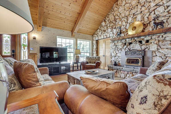 A cozy living room with a stone wall, wooden ceiling, leather sofas, a fireplace, and a television. - 25-Acre Hideaway in Oakhurst: Pet Friendly! (Oakhurst)