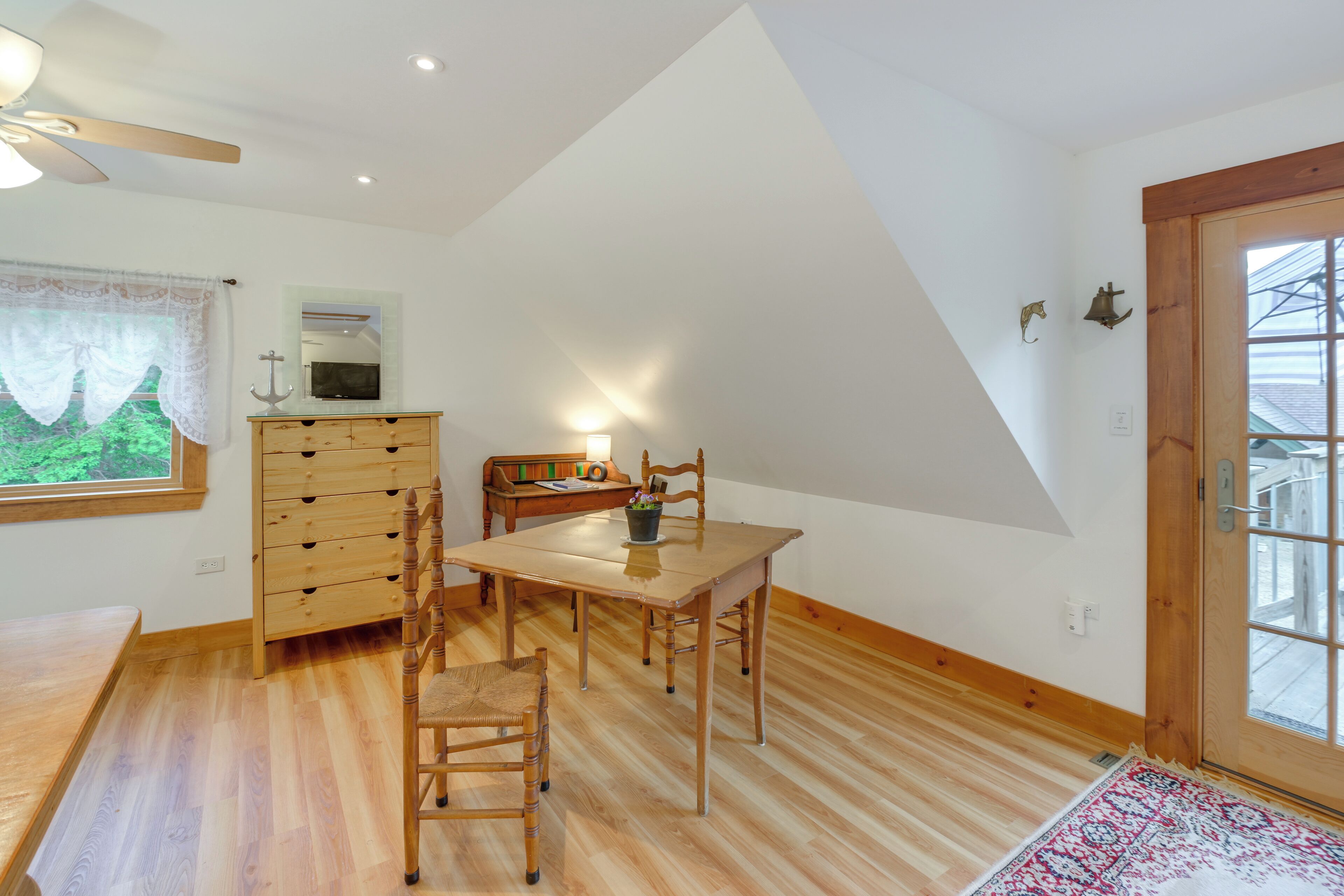 A room with a dining table, chairs, a wooden dresser, a television, and a window with a view of greenery.
