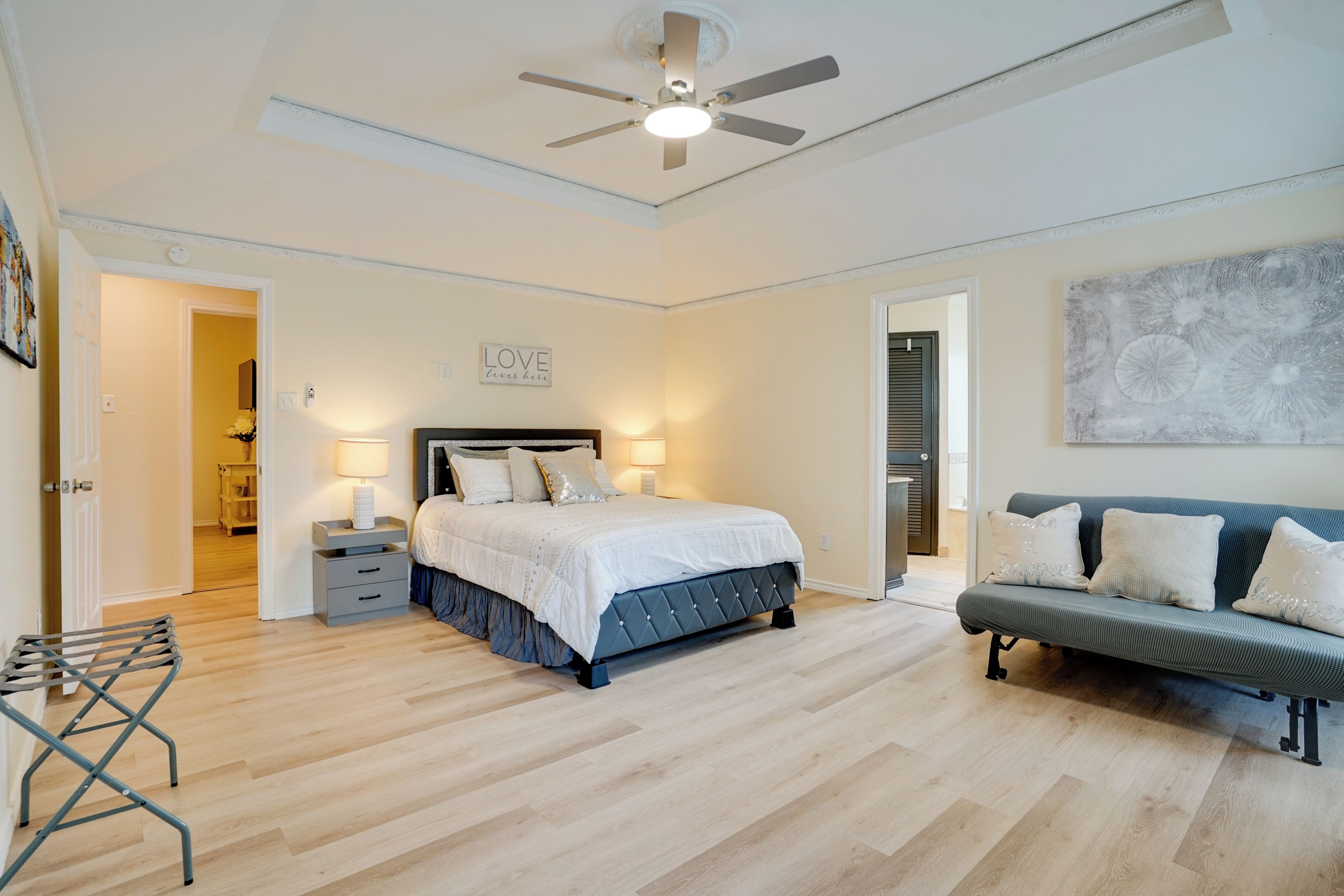 Une chambre à coucher comprenant un lit, des tables de chevet, un ventilateur de plafond, un canapé et un mur agrémenté d’un élément décoratif.