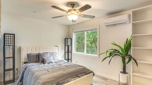 Ein Schlafzimmer mit einem Bett, einem Deckenventilator, einer Klimaanlage, einem Fenster mit Blick auf Bäume und einer Topfpflanze.