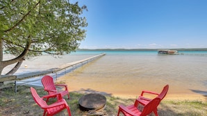 A lakeside setting with two red Adirondack chairs, a fire pit, and a dock extending into the water. - Glen Arbor Home w/ Glen Lake Access & Shared Dock! (Glen Arbor)