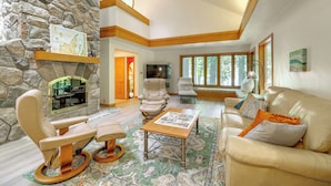 A spacious living room with a stone wall, a fireplace, leather furniture, and large windows. - Glen Arbor Home w/ Glen Lake Access & Shared Dock! (Glen Arbor)