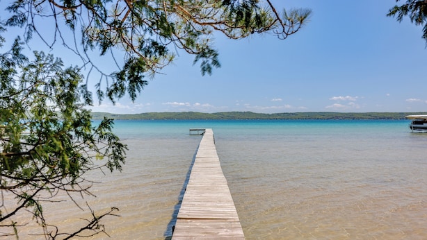 A wooden pier extending into a clear blue body of water, surrounded by trees. - Glen Arbor Home w/ Glen Lake Access & Shared Dock! (Glen Arbor)