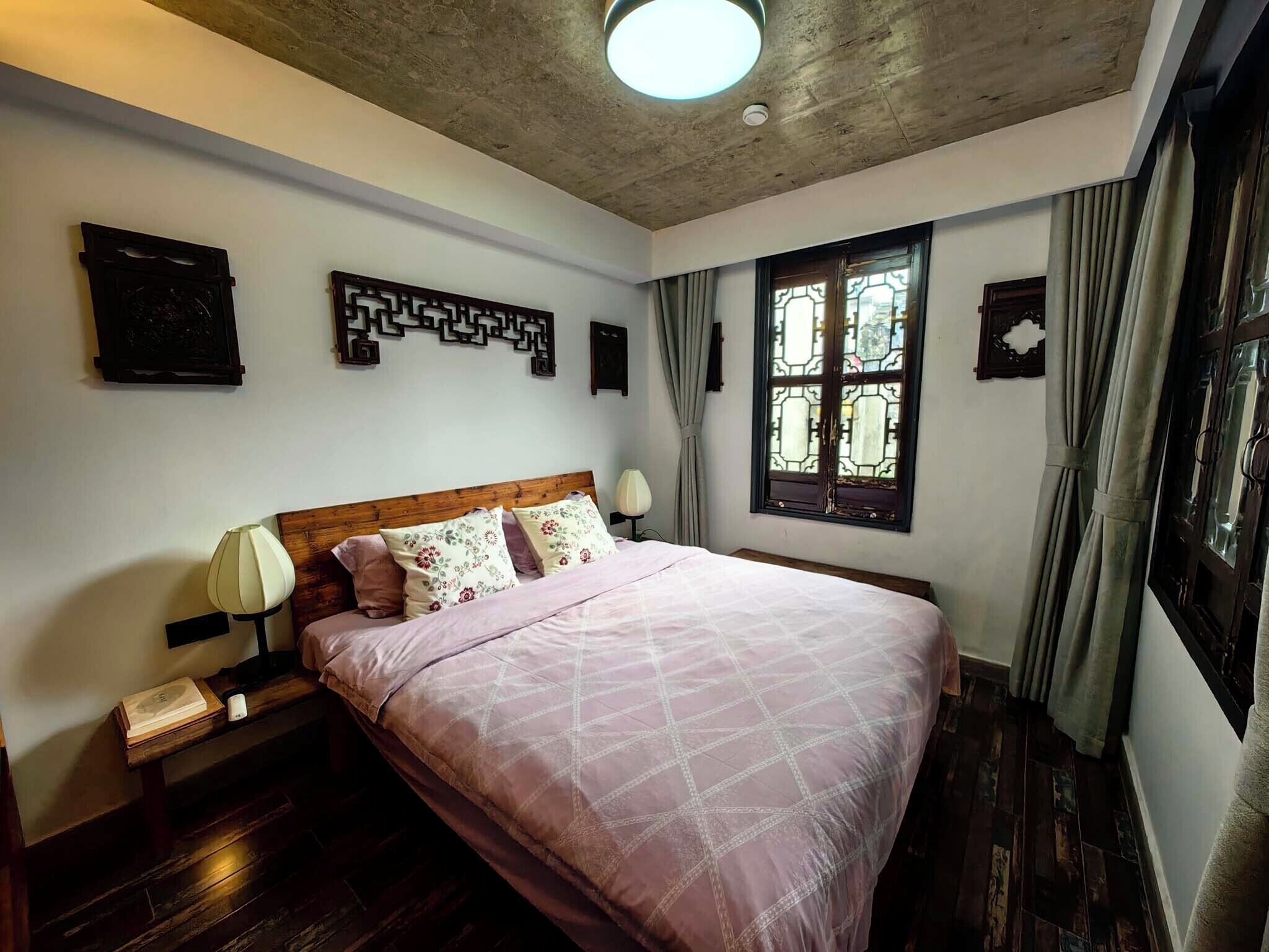 A bedroom with a wooden headboard, a large bed with a pink quilt, two bedside tables with lamps, a window with curtains, and a decorative wall piece.