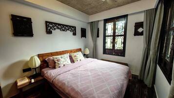 A bedroom with a wooden headboard, a large bed with a pink quilt, two bedside tables with lamps, a window with curtains, and a decorative wall piece.