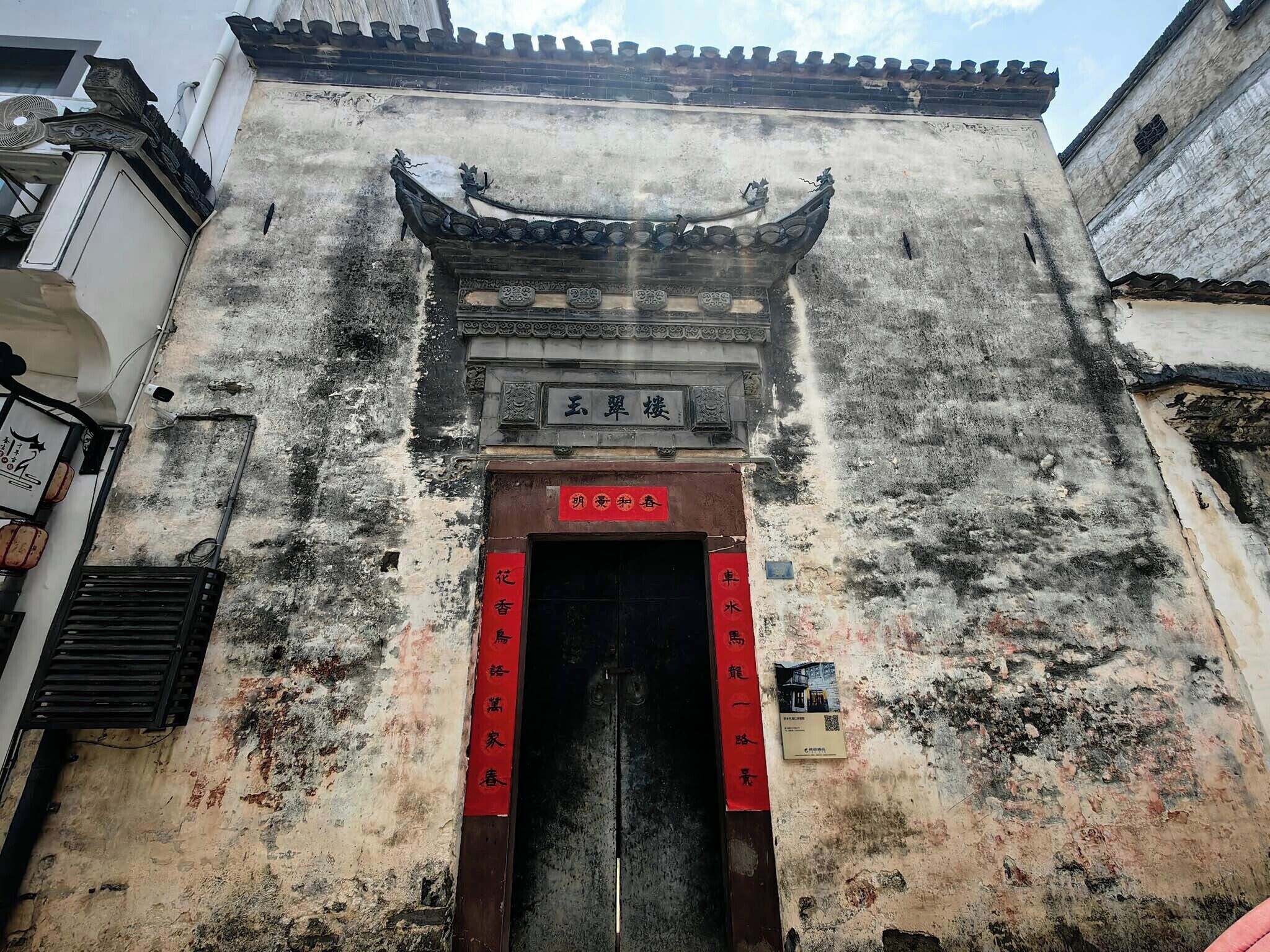 A traditional building with a tiled roof, a central entrance with a sign, and a red banner on the wall.