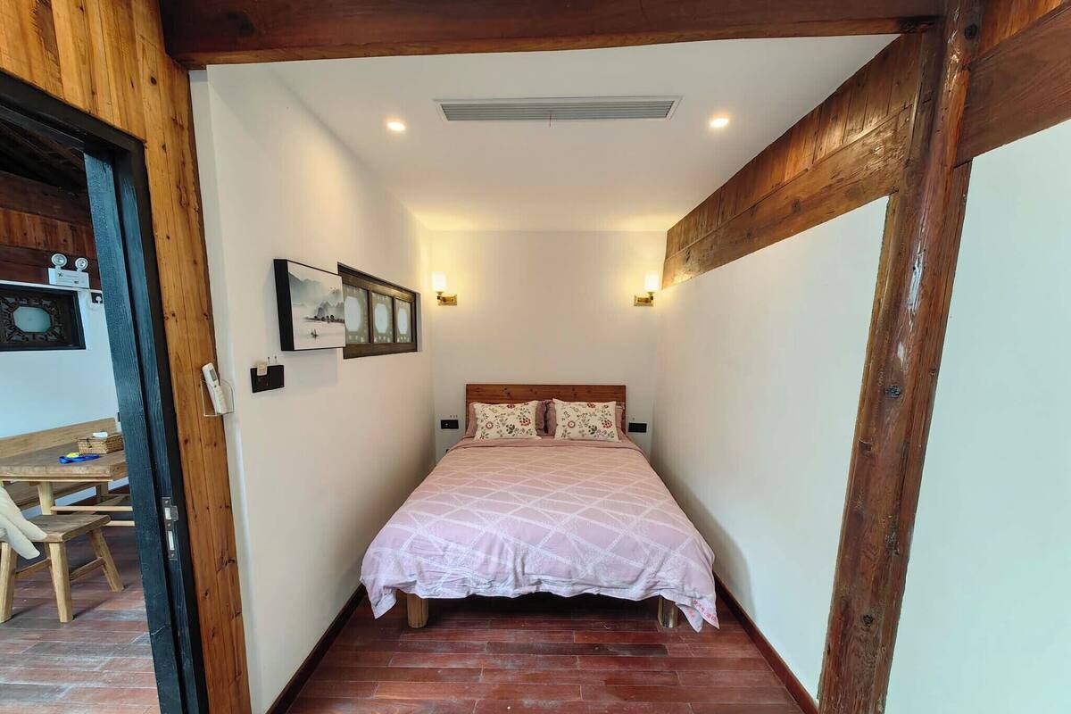 A narrow room with a bed, wooden beams, a small table, and a wall-mounted picture.