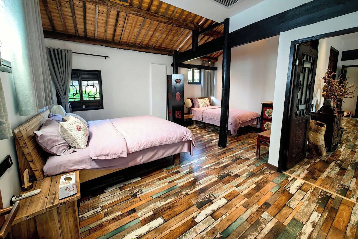 A bedroom with a wooden ceiling, a bed with a pink bedspread, a wooden floor, and a window with curtains.