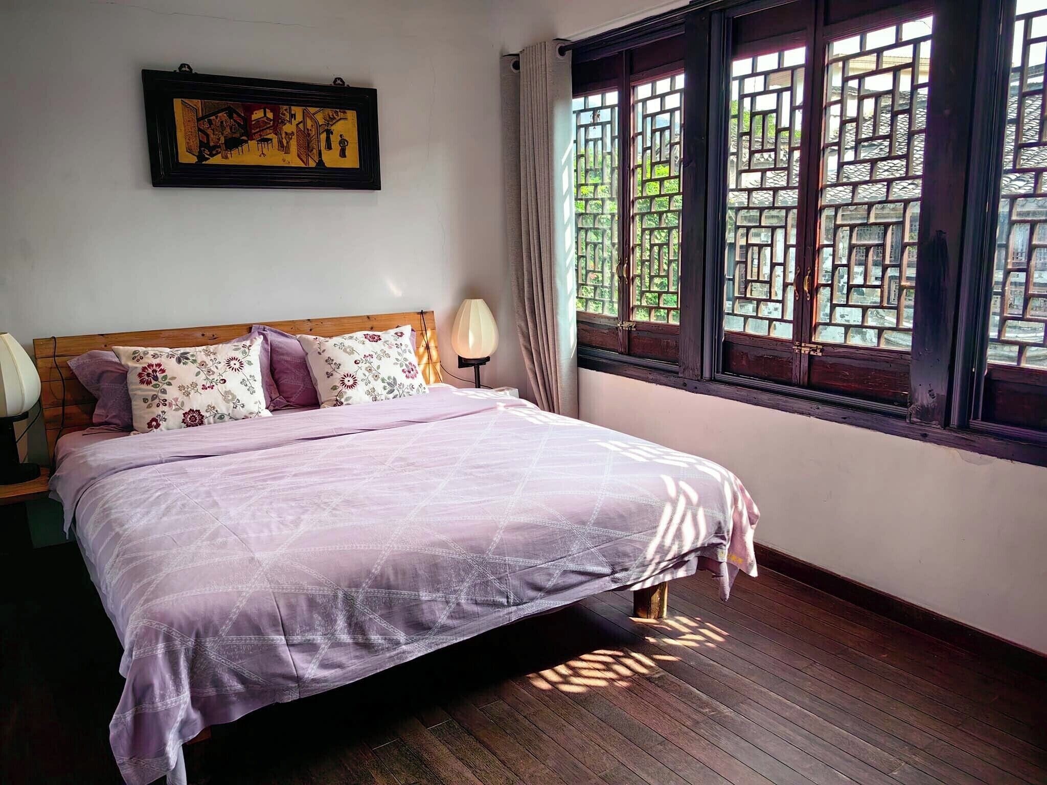 Un dormitorio con una cama, una mesita de noche, una lámpara, una ventana con diseño de celosía y un cuadro en la pared.