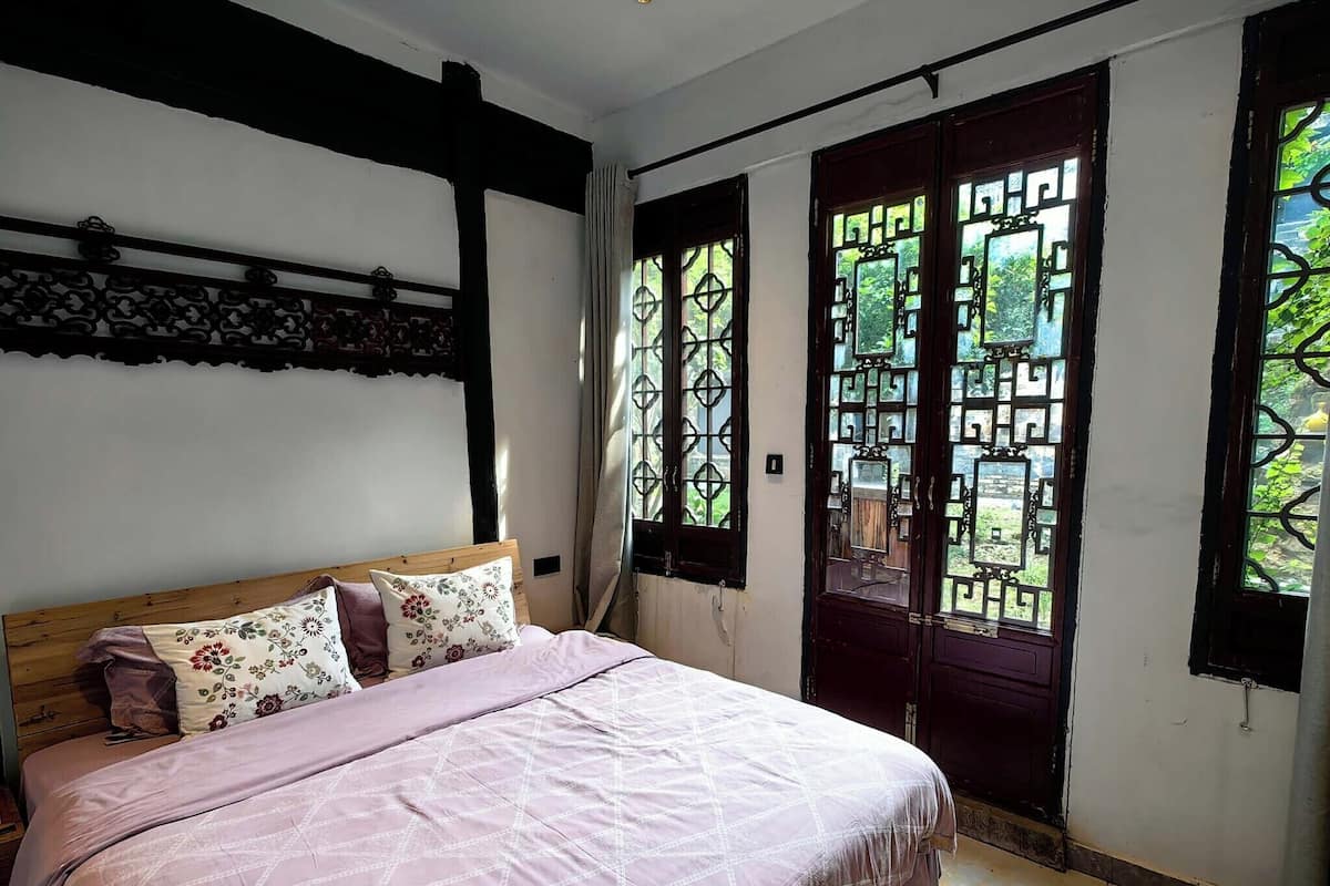 A bedroom with a bed, a wooden headboard, a window with decorative metal grilles, and a view of greenery outside.