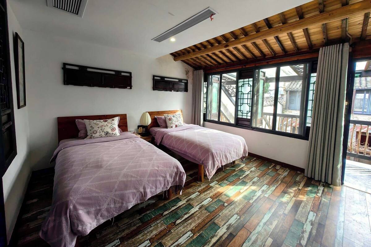 A room with two beds, wooden flooring, a sloped ceiling, and a window with a view of outdoor stairs.