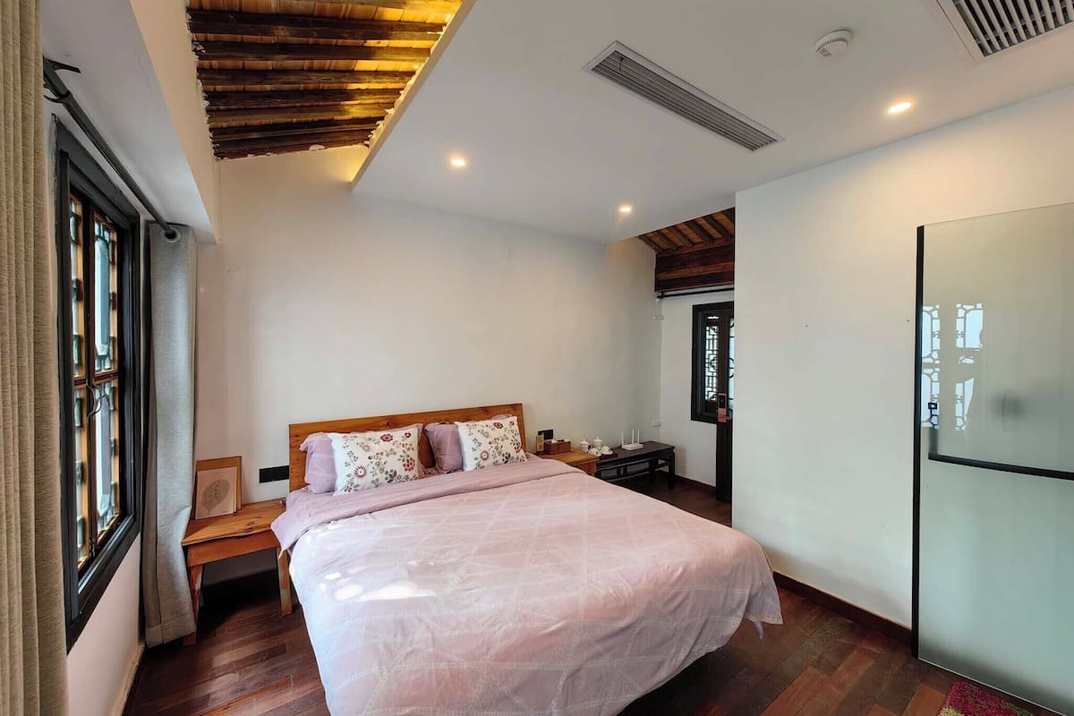 A bedroom with a wooden ceiling, a bed with a floral bedspread, a bedside table, and a window with curtains.