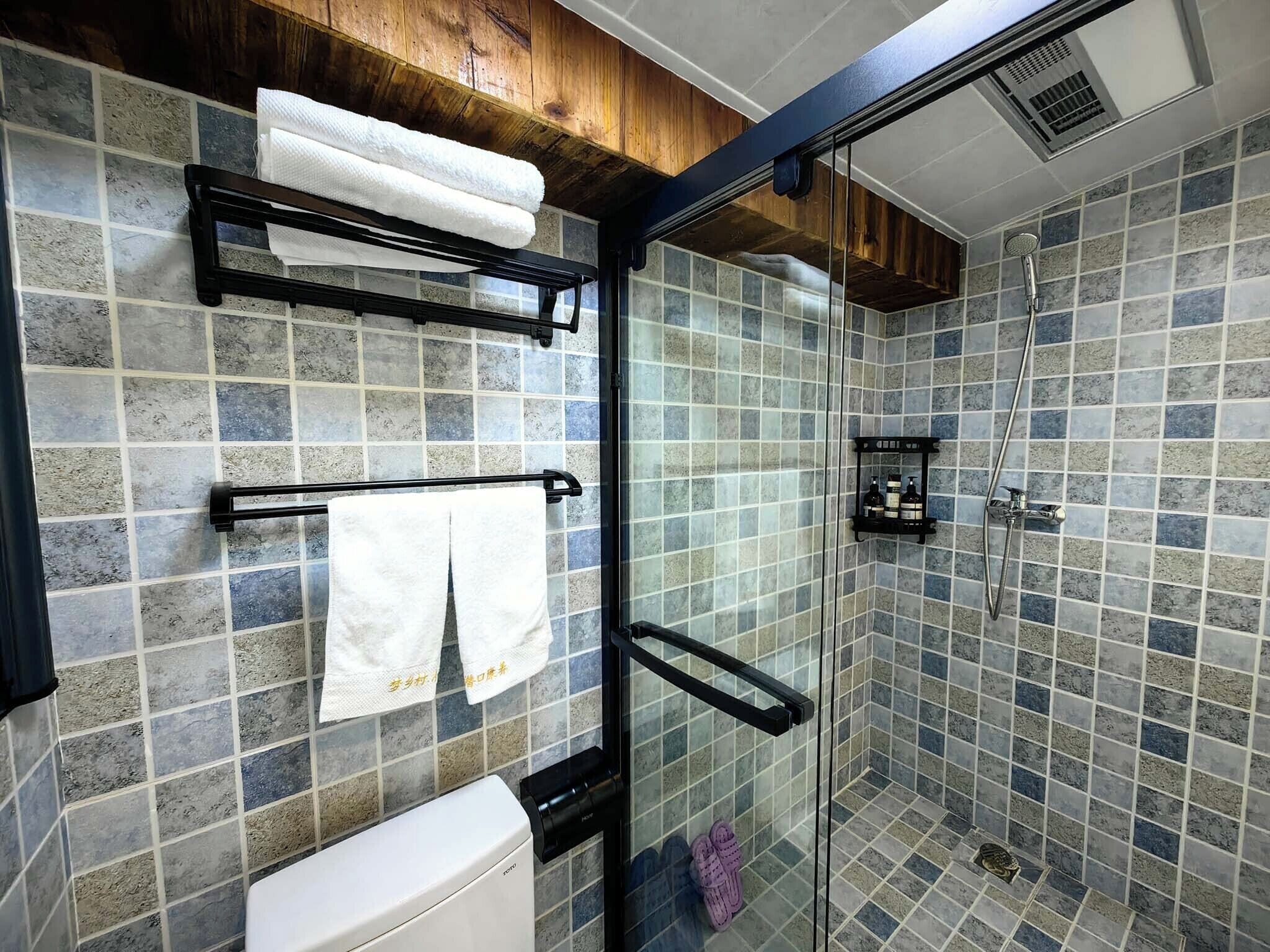A bathroom with a glass shower enclosure, a toilet, a towel rack with towels, and a shelf with toiletries.