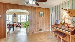 Ein Zimmer mit Holzboden, einer Essecke mit Stühlen und Tisch, einem Klavier, einem Deckenventilator und einer Wand mit einer Gitarre.