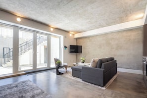 A modern living room with a grey sofa, a flat-screen TV mounted on the wall, and a staircase leading to an outdoor area. - West Van Luxury Retreat 2BR 2BA Oceanview (West Vancouver)